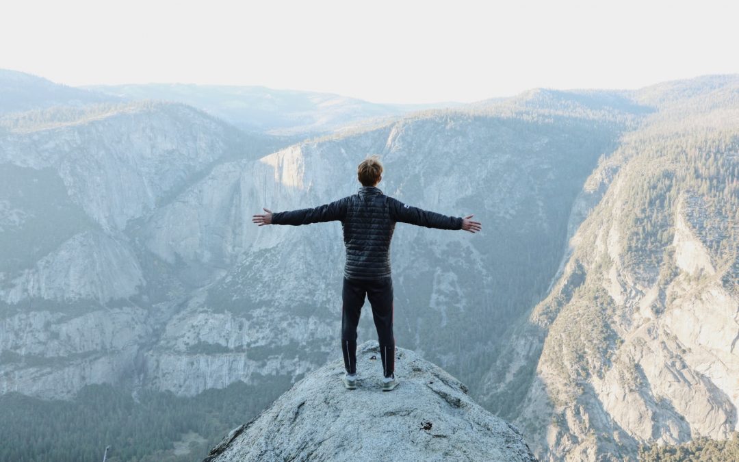 man opening his arms wide open on snow covered cliff with view of mountains during daytime